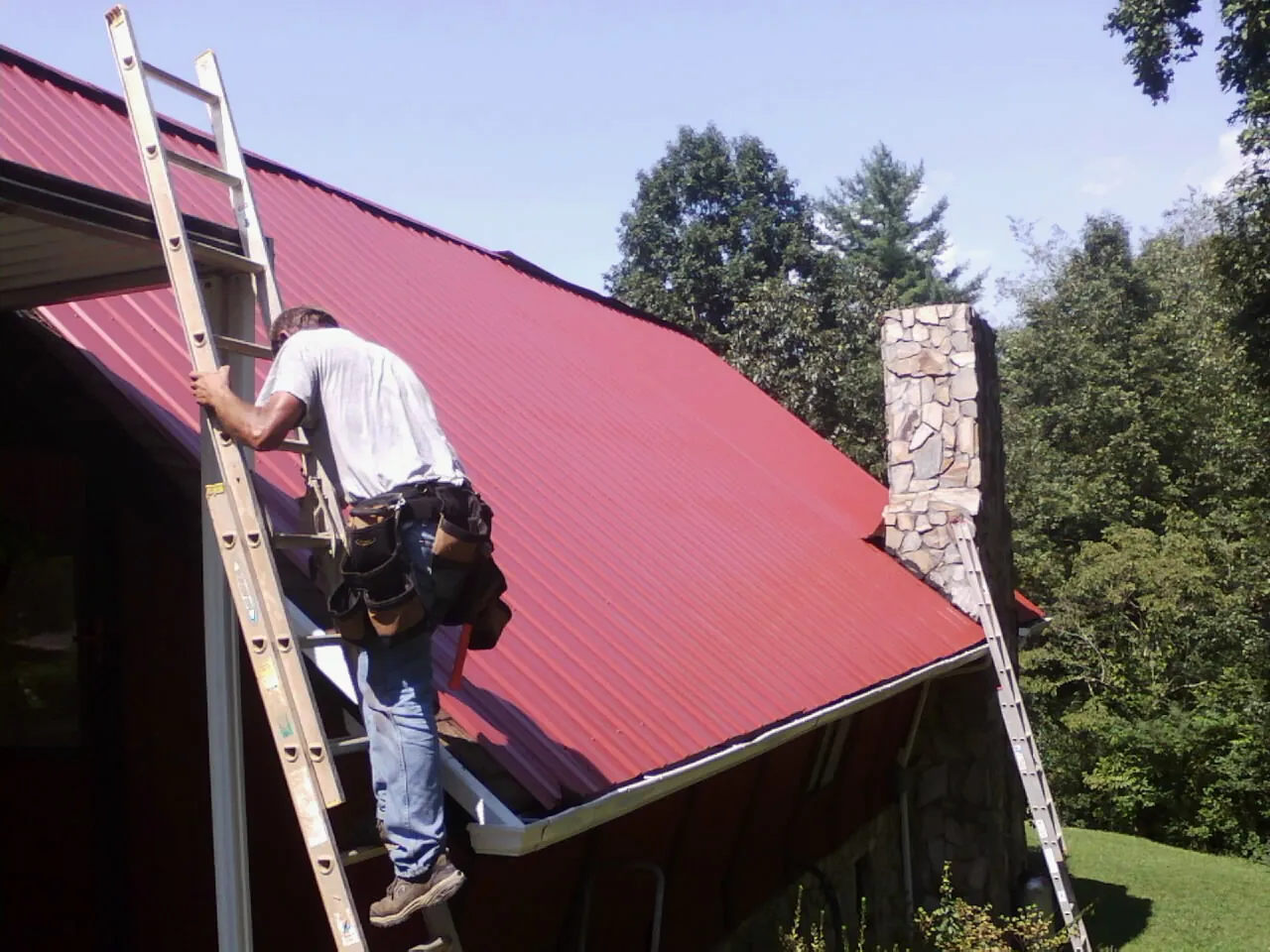 Roofer on a ladder beside a red metal roof and stone chimney — Soffit & Fascia Repair by Roofing Brothers in Walnut Bottom